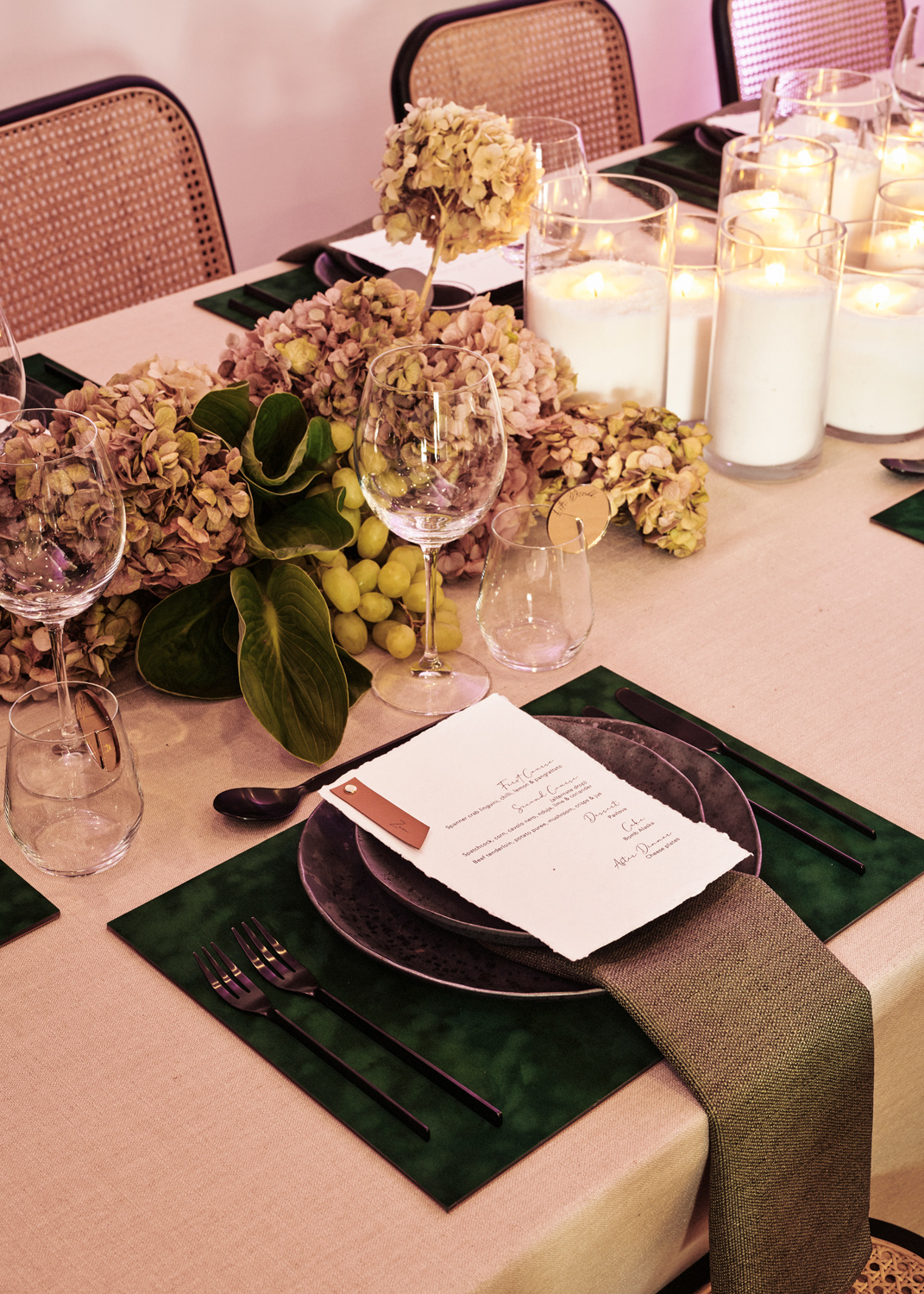 Stylish table setting featuring rattan chairs, beige linen table cloth, moss coloured napkins, printed menus, green and purple hydrangeas and candles.