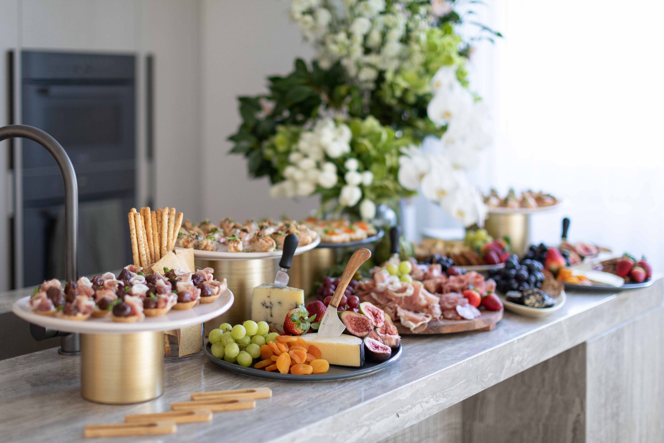 Grazing table set up on a kitchen bench adorned with finger food, cheeses, crackers, fruits and antipasto, and fresh florals