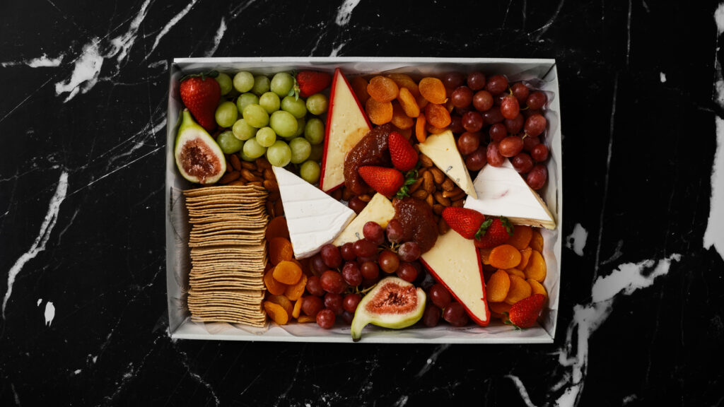 Artisan cheese box artfully assembled by Boxed by Blonde Butler for Christmas. Several gourmet cheeses with fresh and dried fruit and crackers are beautifully displayed.